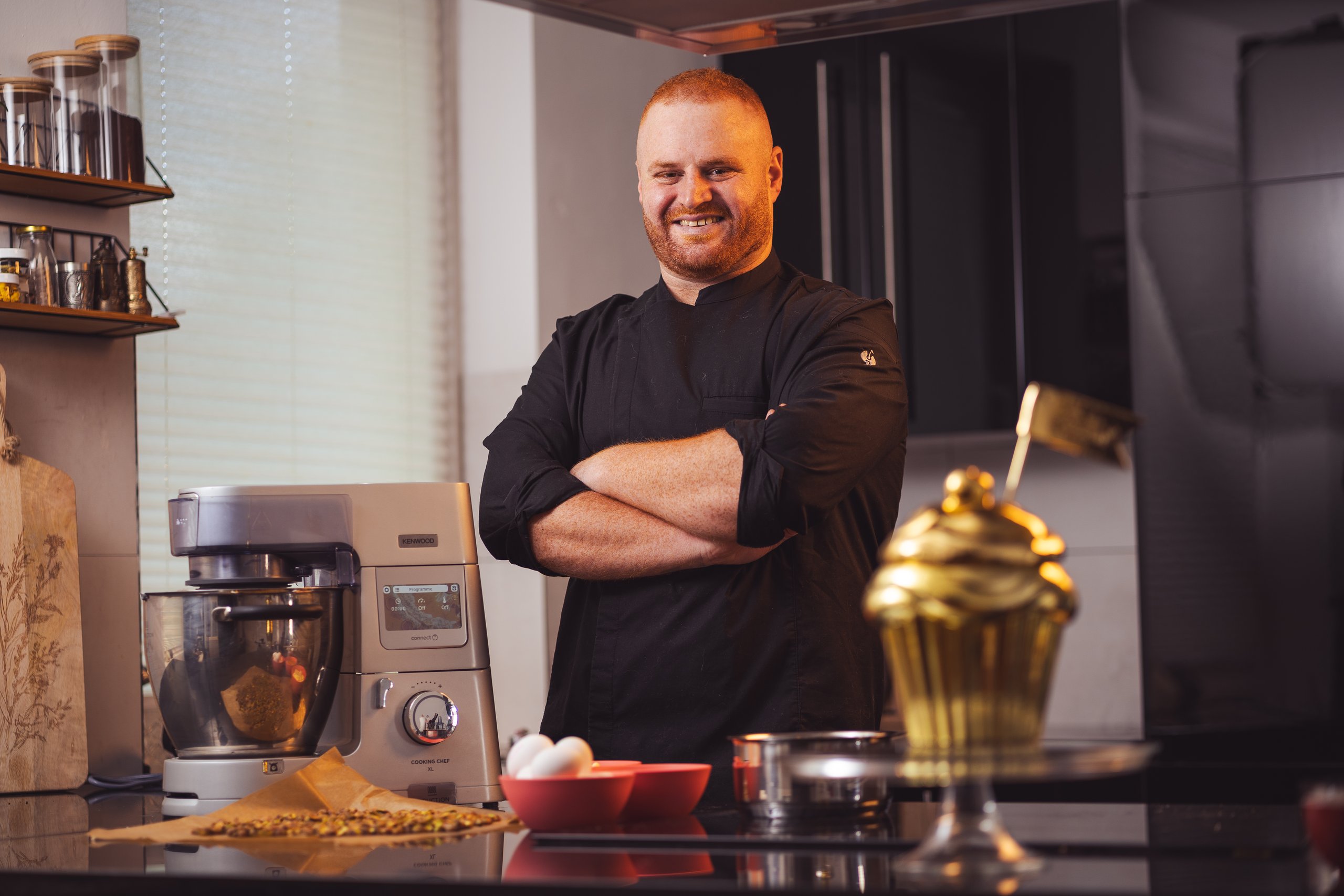 Raheem Haidar mit verschränkten Armen in seiner Küche, neben ihm ein goldener Cupcake, Foodfotografie mit Blitzlicht