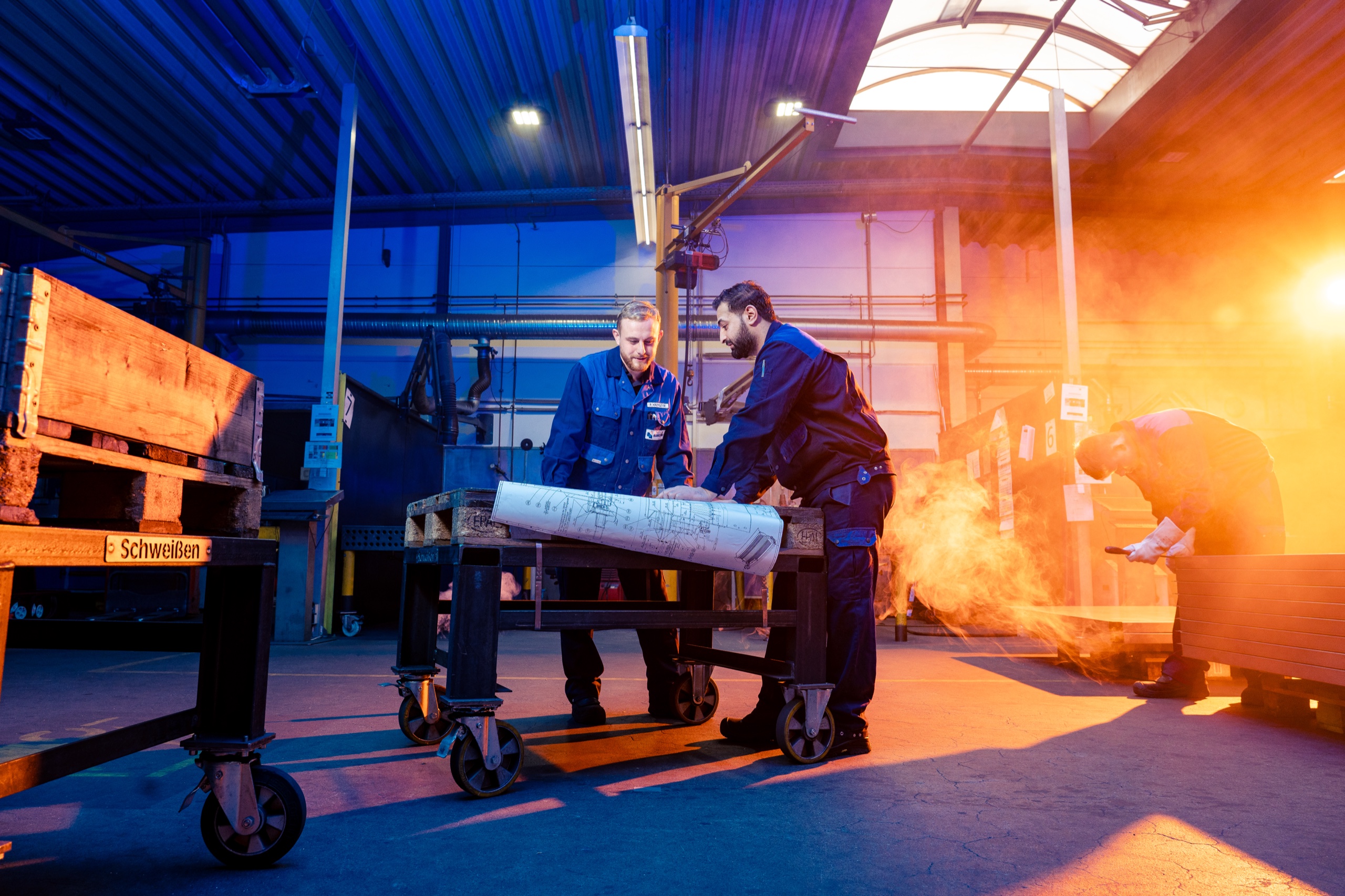 Industriefotografie Hannover: Zwei Mitarbeiter von Blekotec besprechen einen Plan in der Werkhalle, dahinter wird geschweißt