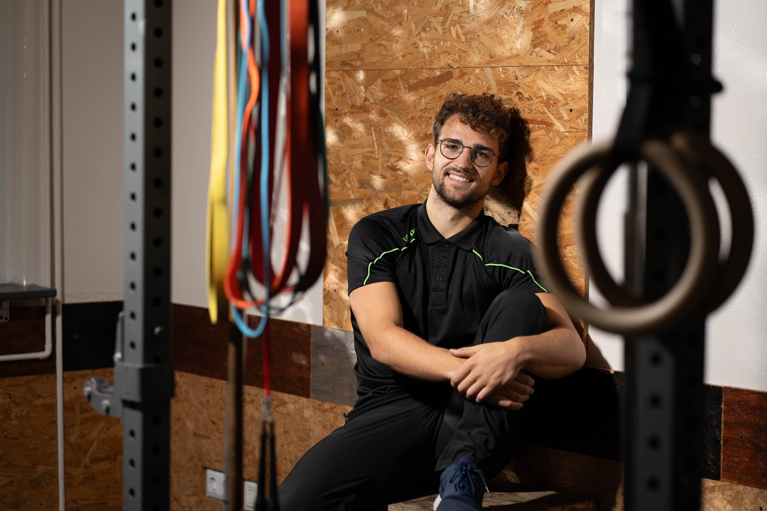 Personal Trainer Lukas Kazimierski sitzt lächelnd im Trainingsraum, im Vordergrund Turnringe